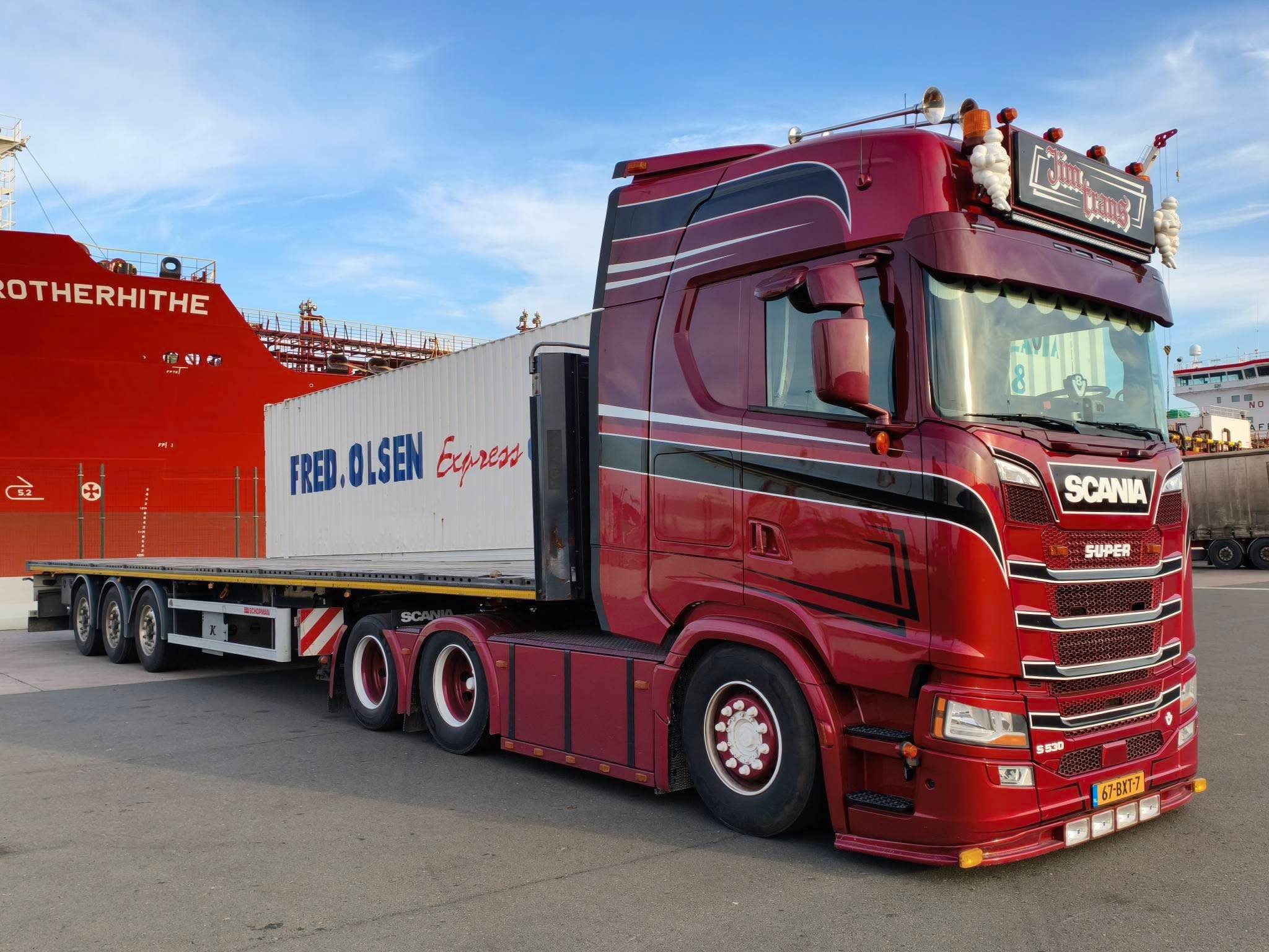Rode Scania vrachtwagen met open oplegger en lading bij een haven