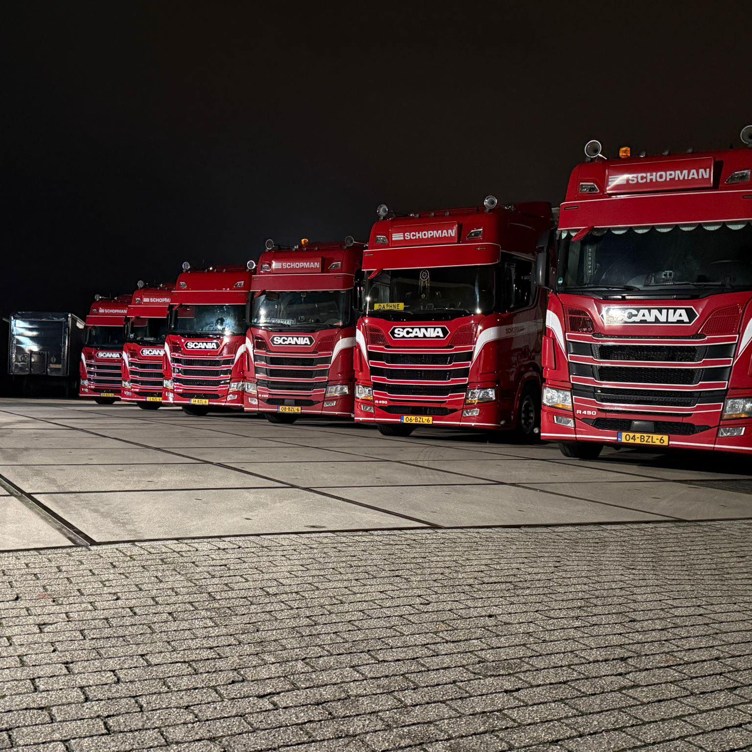 Rij rode Scania vrachtwagens van Schopman Transport opgesteld op een terrein in de avond